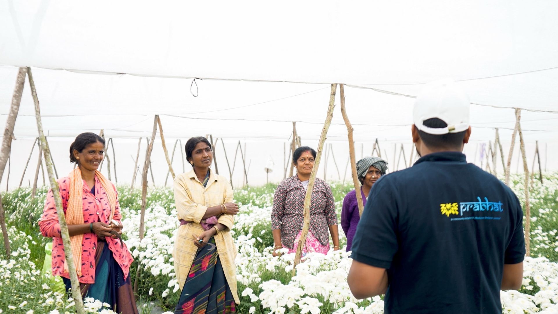 Field officer speaks with women farmers standing among white flowers in a covered greenhouse