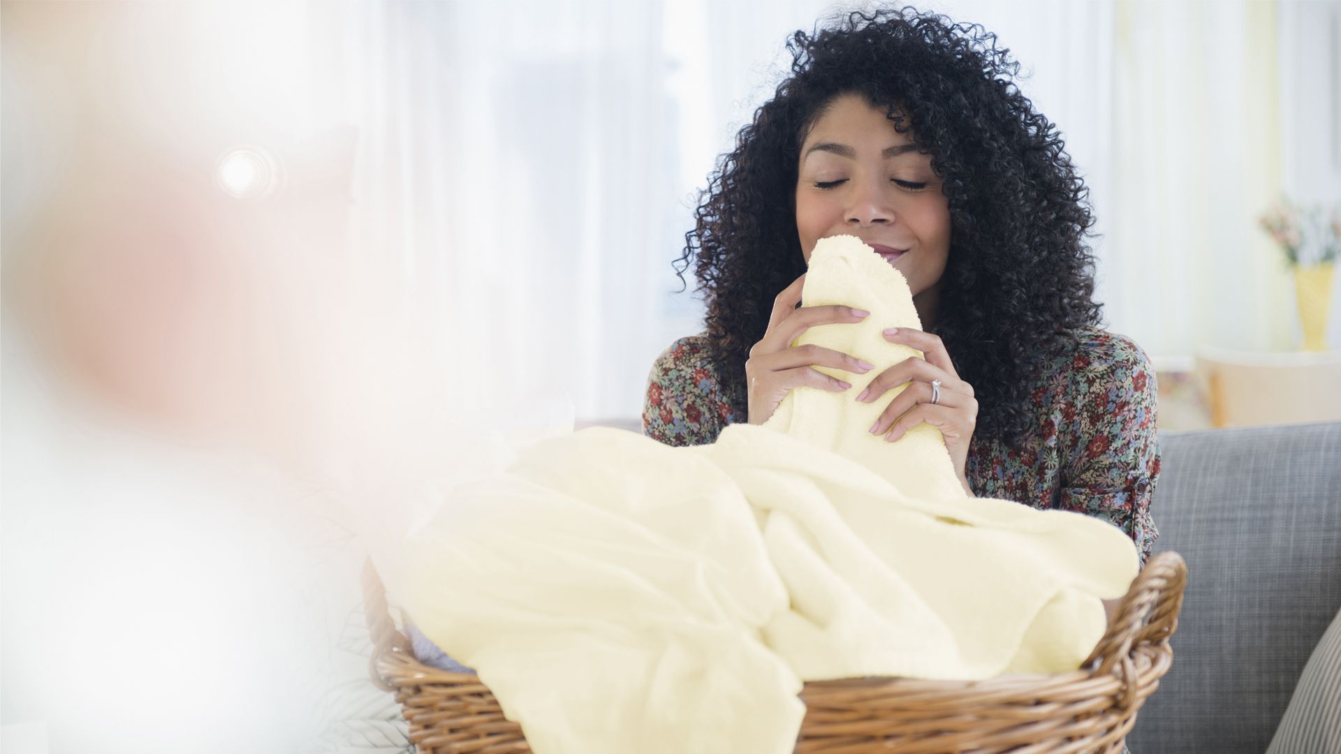 A person with curly hair, holding a large yellow towel near their face, smelling the fragrance, while sitting behind a full wicker basket. The background features a bright room with light curtains and flowers on the right.