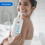 Person holding Dove Exfoliate Away body wash bottle near shoulder in a bright bathroom setting. The image is tagged ‘Geography’.