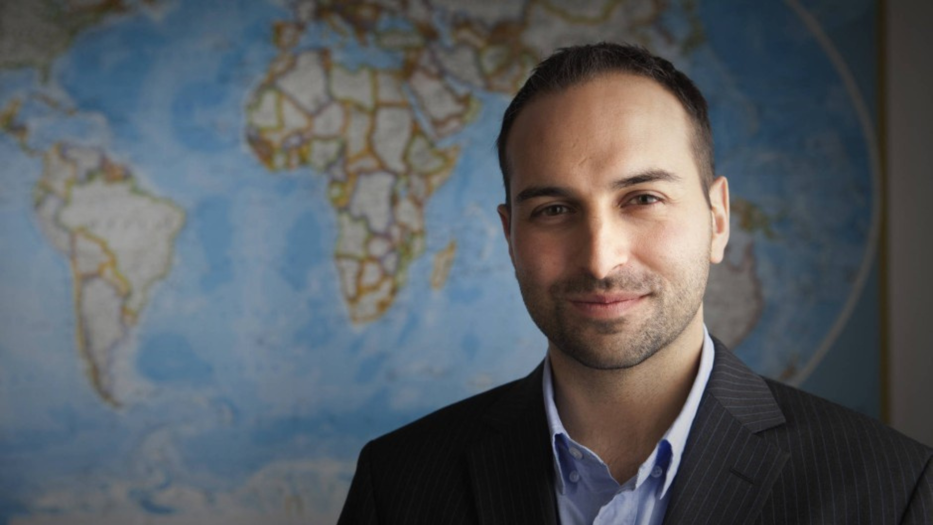 A headshot of Ricken Patel. Ricken has a large world map poster behind him. He is wearing a light blue shirt and a suit jacket, as he looks towards the camera.