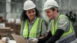 Two people in hard hats in a factory chatting and looking at a laptop
