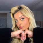 Person with long blonde hair wearing a black fuzzy garment, hands clasped under chin displaying a large ring and manicured nails.