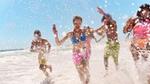 Five people running through shallow water at a beach, splashing waves around them under a clear blue sky. They are dressed in colourful swimwear and appear to be enjoying a sunny day.