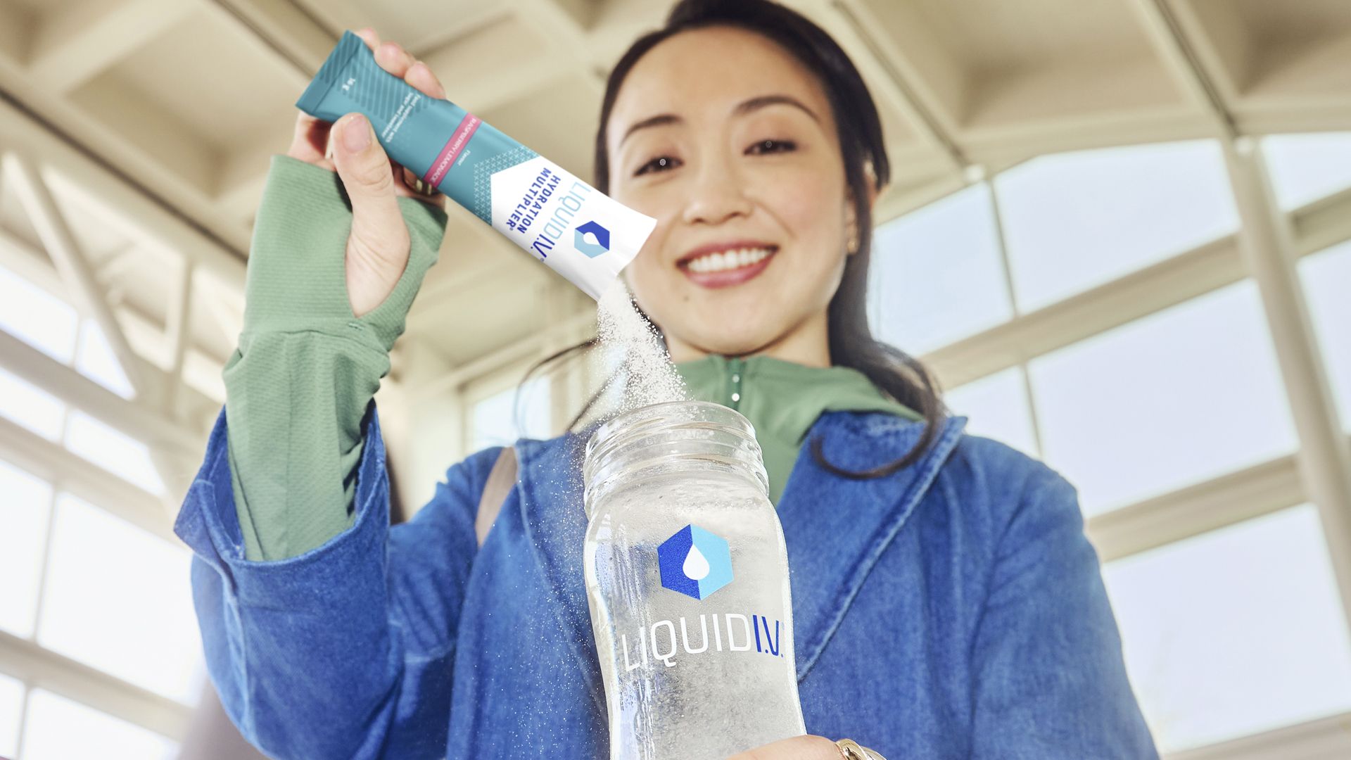 A person in a blue jacket with green sleeves is pouring a sachet of Liquid I.V. into a clear bottle with the Liquid I.V. logo on it. The scene is set in a bright room with large windows. This image captures a moment of preparing a hydration supplement, perfect for health and wellness enthusiasts.
