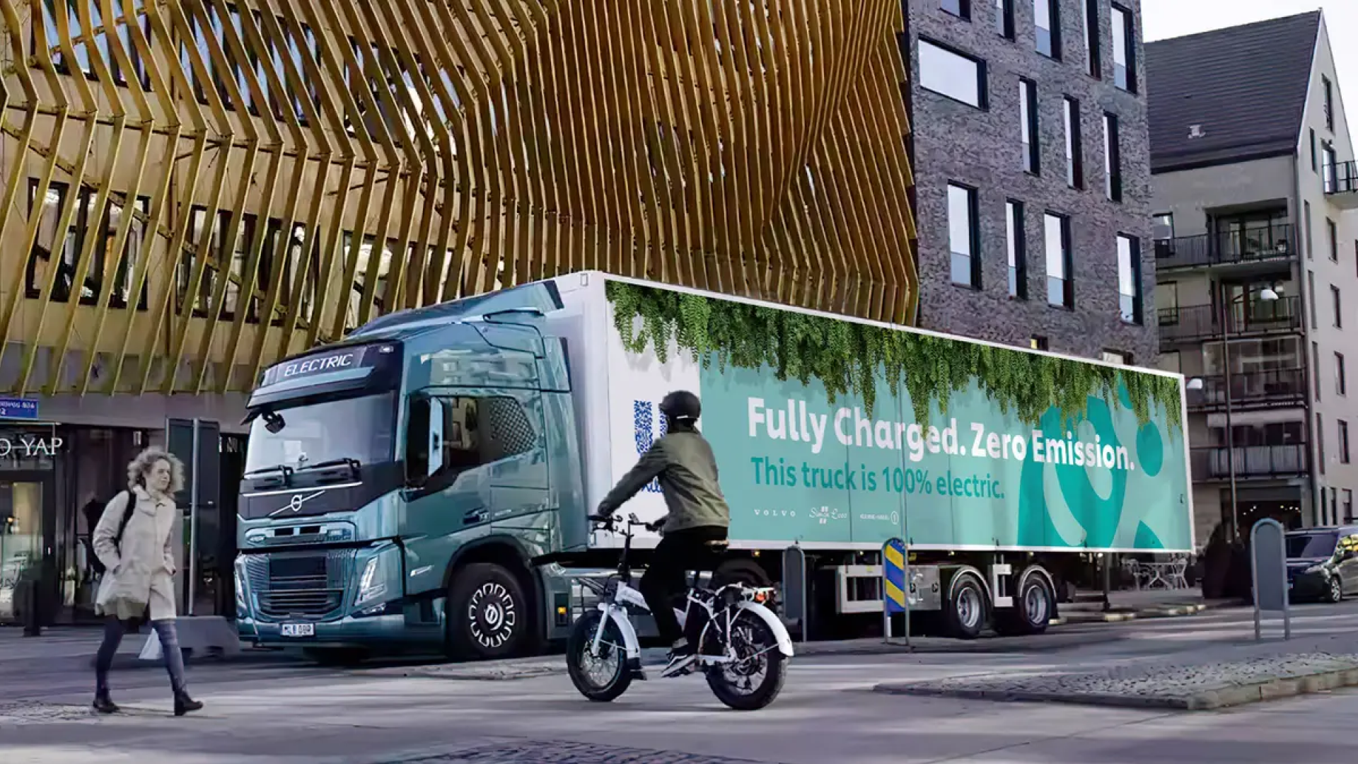 An electric truck with 'Fully Charged. Zero Emission. This truck is 100% electric.' written on its side, driving through a modern urban area with a cyclist and a pedestrian nearby, and contemporary buildings in the background.