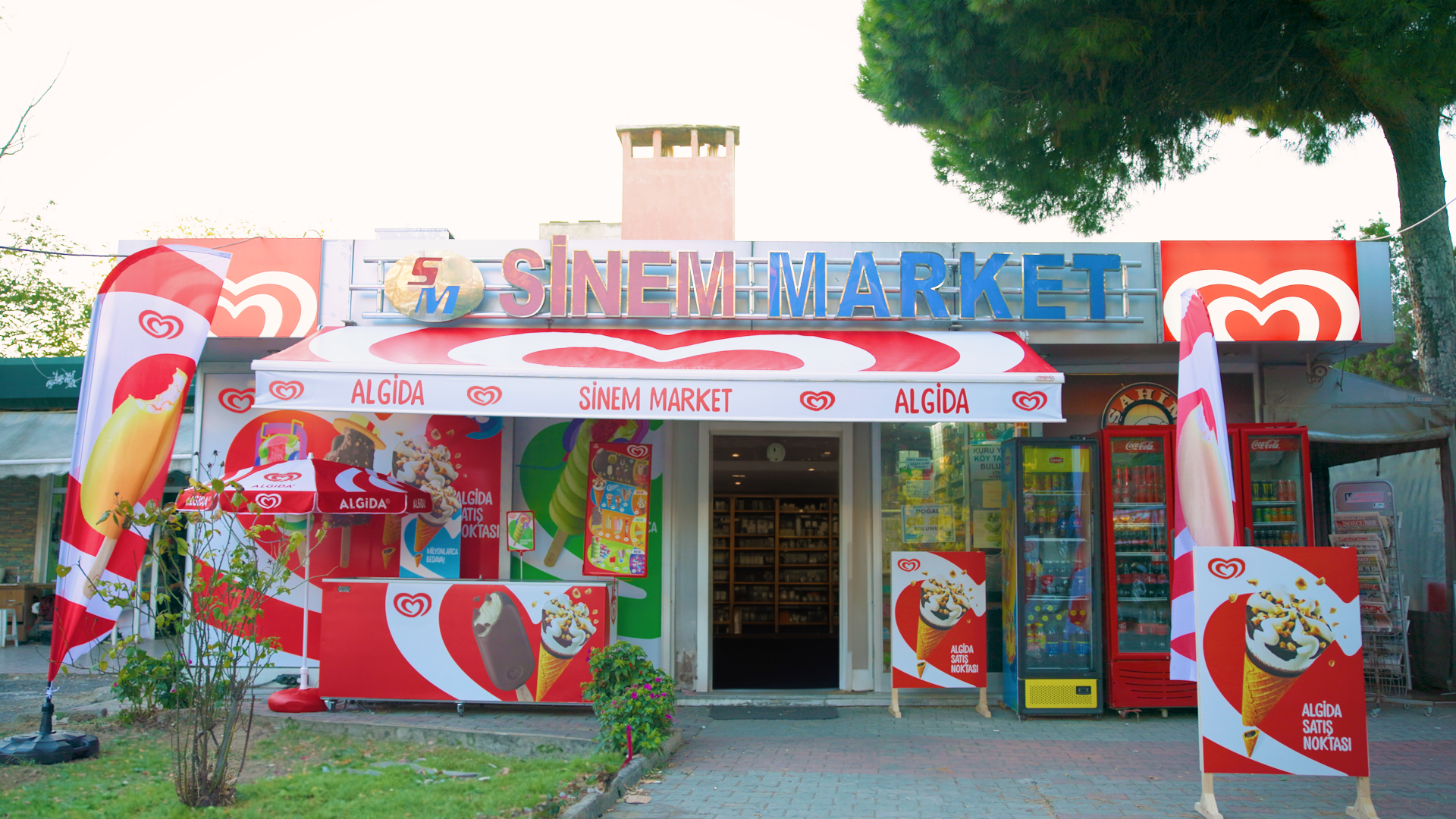 External view of a mini market displaying Wall’s (Algida) branding on the sign, awning, flags and umbrellas.