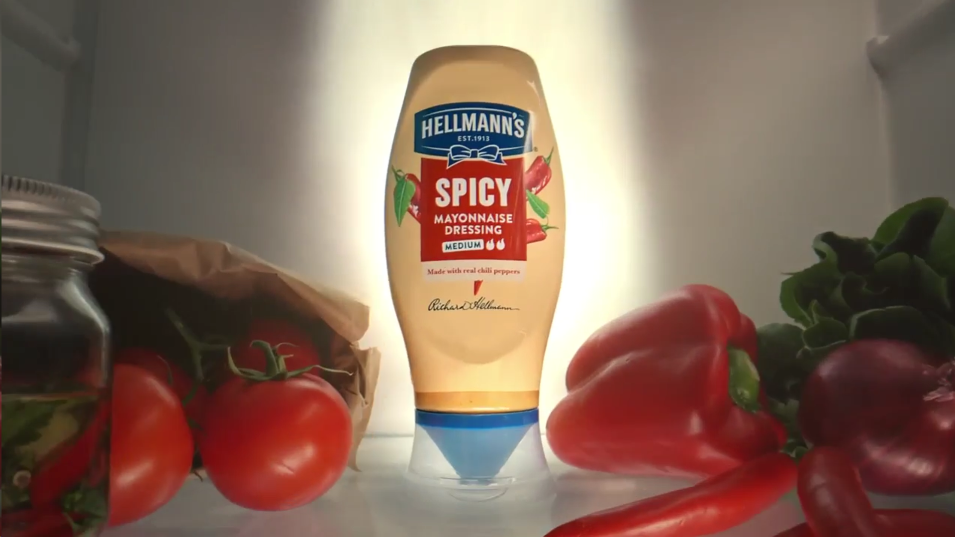A glowing bottle of Hellmann’s Spicy Mayonnaise in a fridge surrounded by different vegetables.
