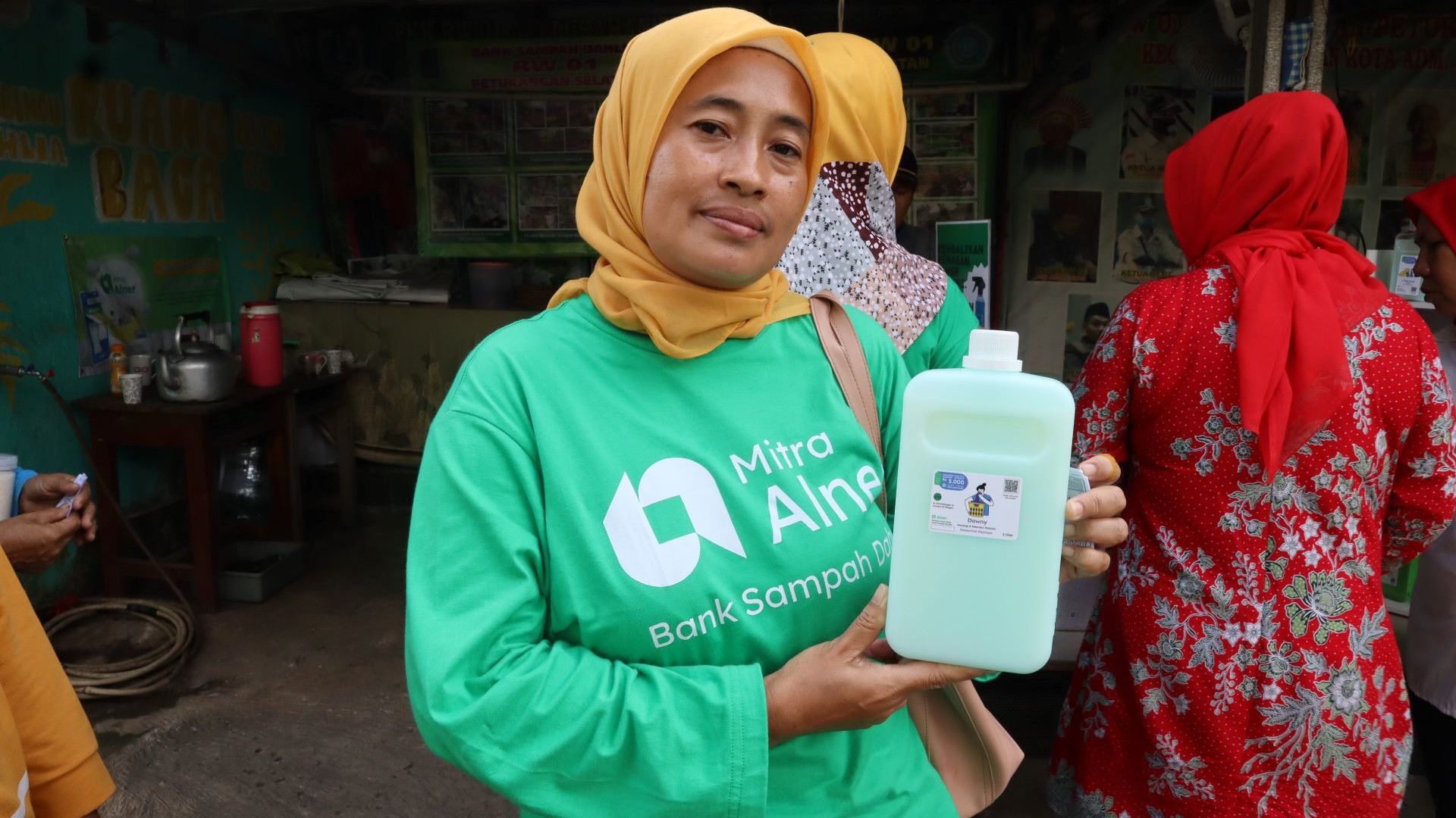 Woman wearing long sleeved green t-shirt and yellow head scarf smiling at the camera holding a large bottle of green liquid product.