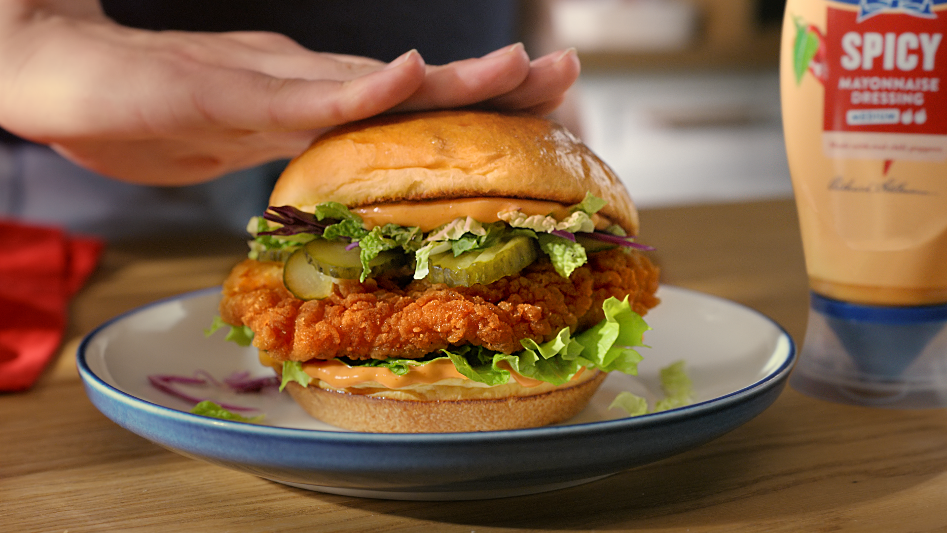 A chicken burger on a plate on a wooden table. To the right of the plate is a bottle of Hellmann’s spicy mayo. A hand is pressing down lightly on top of the burger.