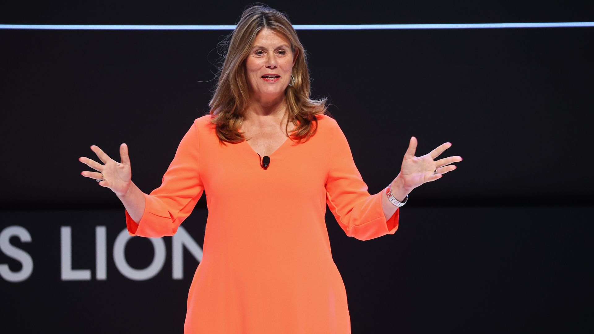 Conny Braams in oragnge dress with arms extended speaking on stage