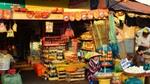 A vibrant outdoor market stall showcasing a variety of goods for sale. The stall is stocked with colourful products, neatly displayed on shelves and in baskets. In the foreground is a woman wearing a traditional conical hat and a striped shirt sitting next to a basket of goods.