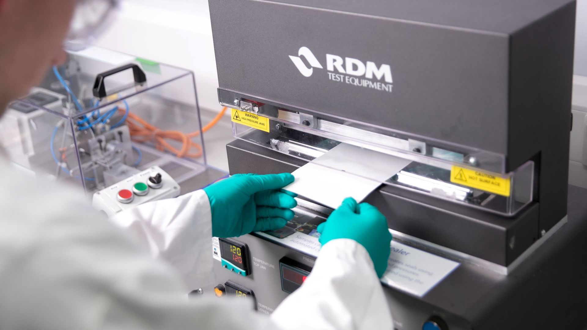 A laboratory technician wearing green gloves places a sample sheet into an RDM Test Equipment machine. The background shows additional laboratory apparatus and tools.