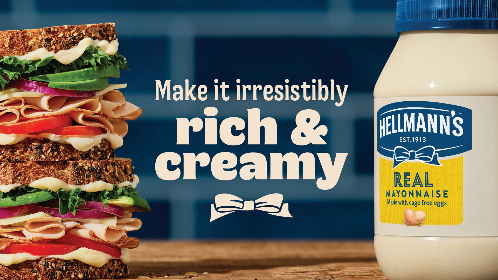 On the left is a multi-layered sandwich stacked with avocado, lettuce, onion, tomato, cheese, deli meats and bread, with visible layers of mayonnaise. To the right, is a jar of Hellmann’s Real Mayonnaise. Large text in the centre reads: “Make it irresistibly rich & creamy” with a bow icon below the text.
