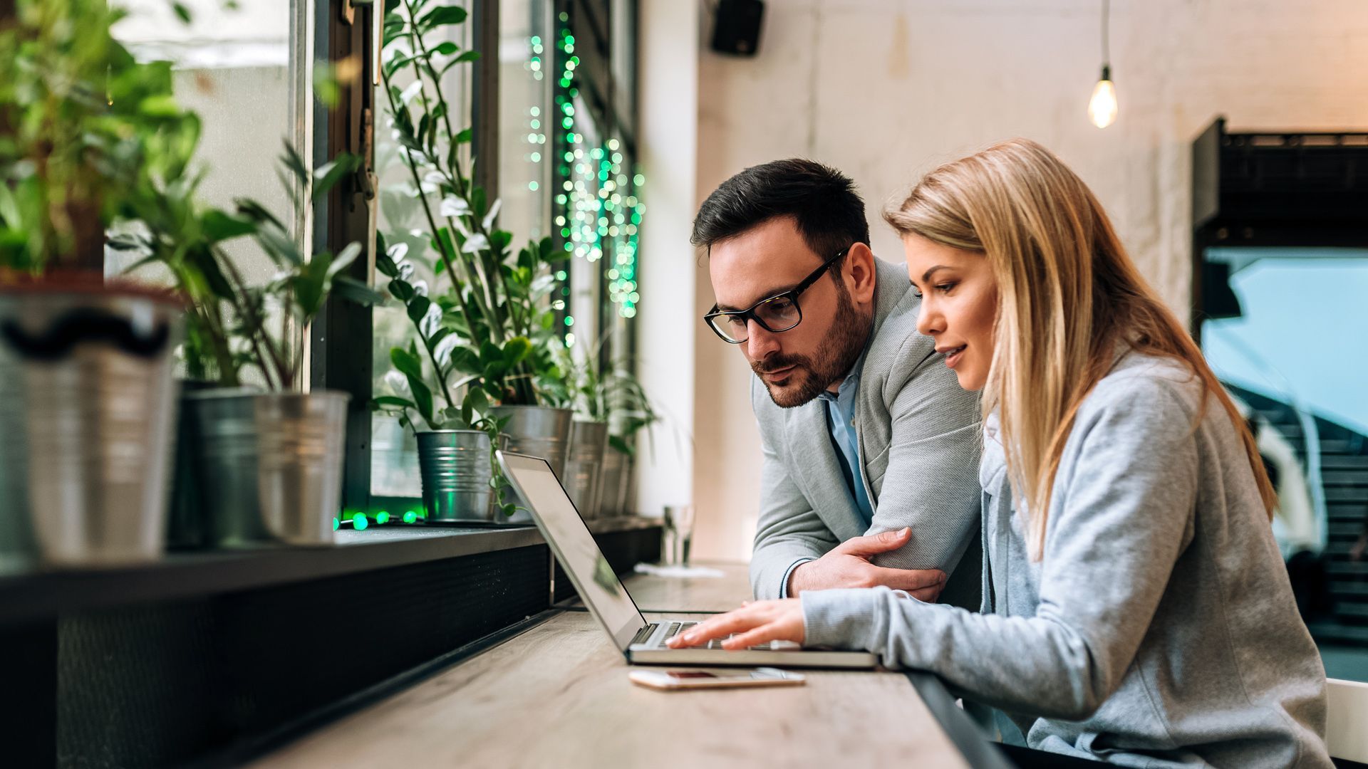 A man and a woman sitting together, looking at a laptop screen, with green plants in the background.