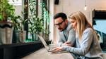 A man and a woman sitting together, looking at a laptop screen, with green plants in the background.