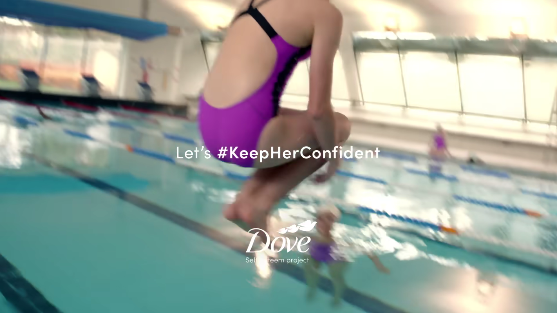 A young girl dive bombing into a 50m swimming pool. She is wearing a purple swim suite.  The text on the screen reads. 'Let’s #keepHerConfident'. At the bottom of the screen is the Dove self esteem project logo.