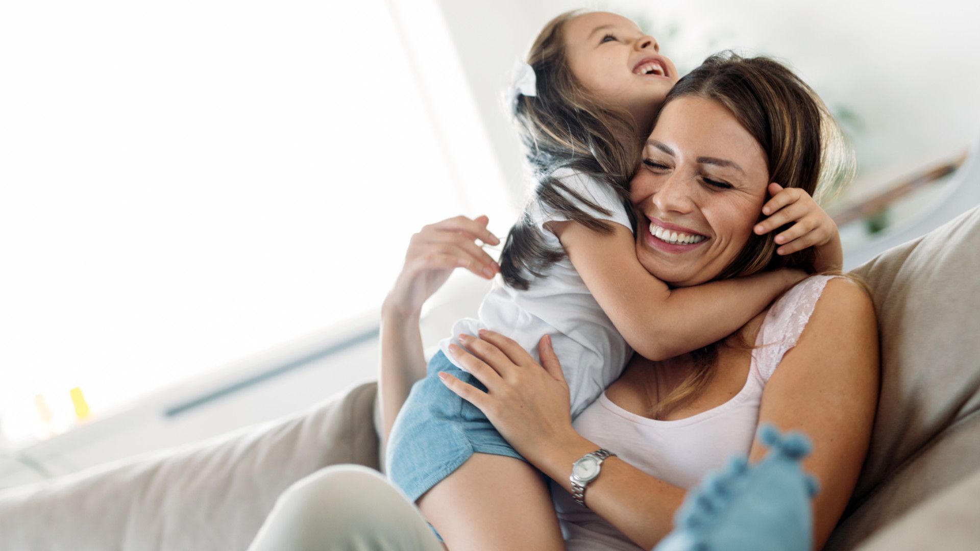 A woman laughing on a couch while a young girl hugs her tightly