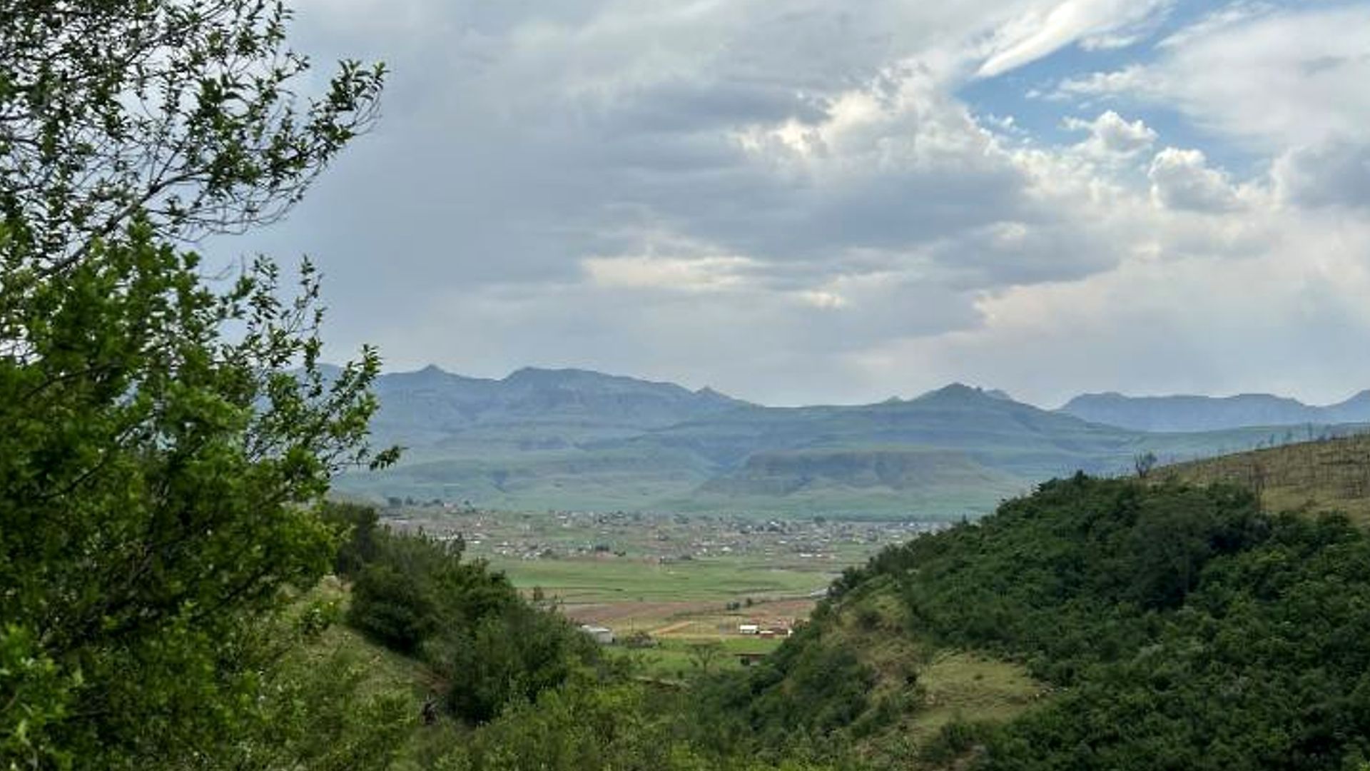 Green hills frame a distant valley town beneath layered blue mountains and a cloudy sky.