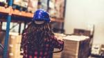 The back of the head of a young woman in a hard hat surveying a warehouse of goods