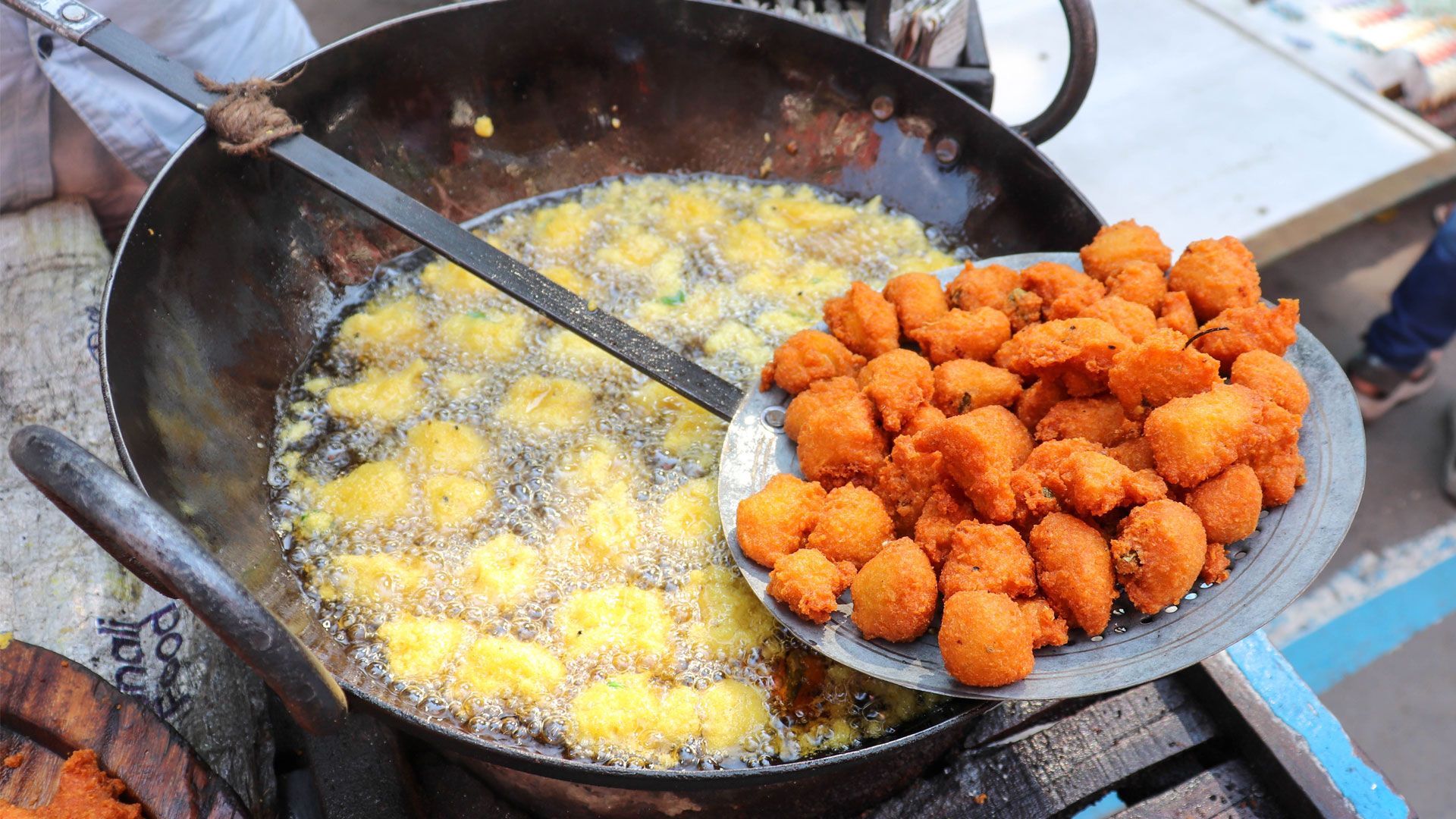 Street food fritters frying in a large pan of bubbling oil, with freshly cooked fritters resting on a slotted spoon.