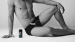 A bare-chested man in black pants sits on the floor in a relaxed pose, one knee raised with his wrist resting on it. A can of Lynx body spray is next to him. A “SCRATCH & SNIFF” label points to his shorts. The image is in black and white.