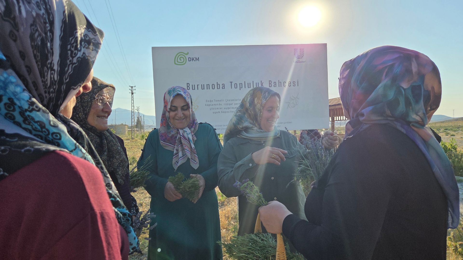 Women in headscarves smile and hold herbs together in a sunny community garden.