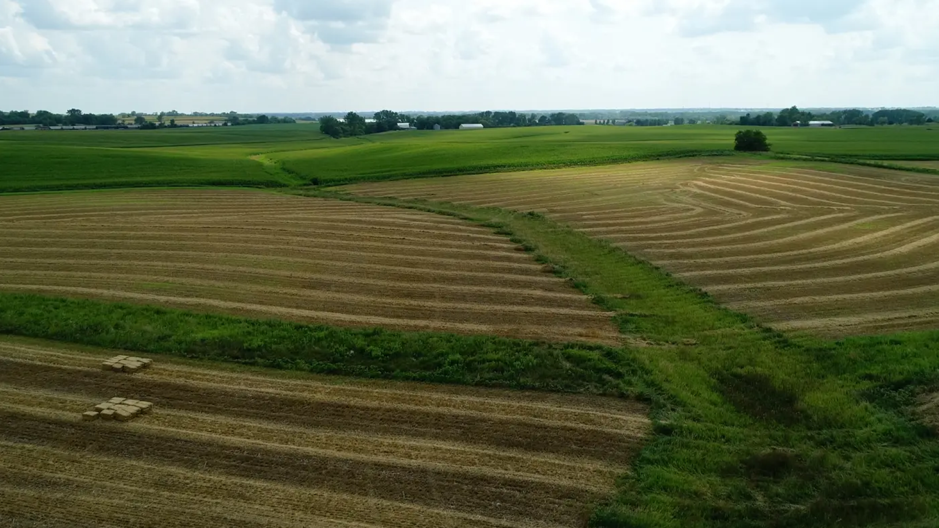 Countryside aerial view showing neatly mowed fields with hay bales and large areas of green grass, under a partly cloudy sky.