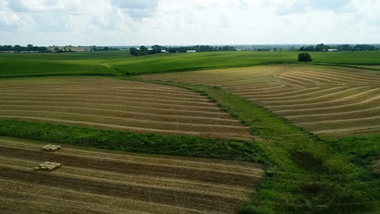 Countryside aerial view showing neatly mowed fields with hay bales and large areas of green grass, under a partly cloudy sky.