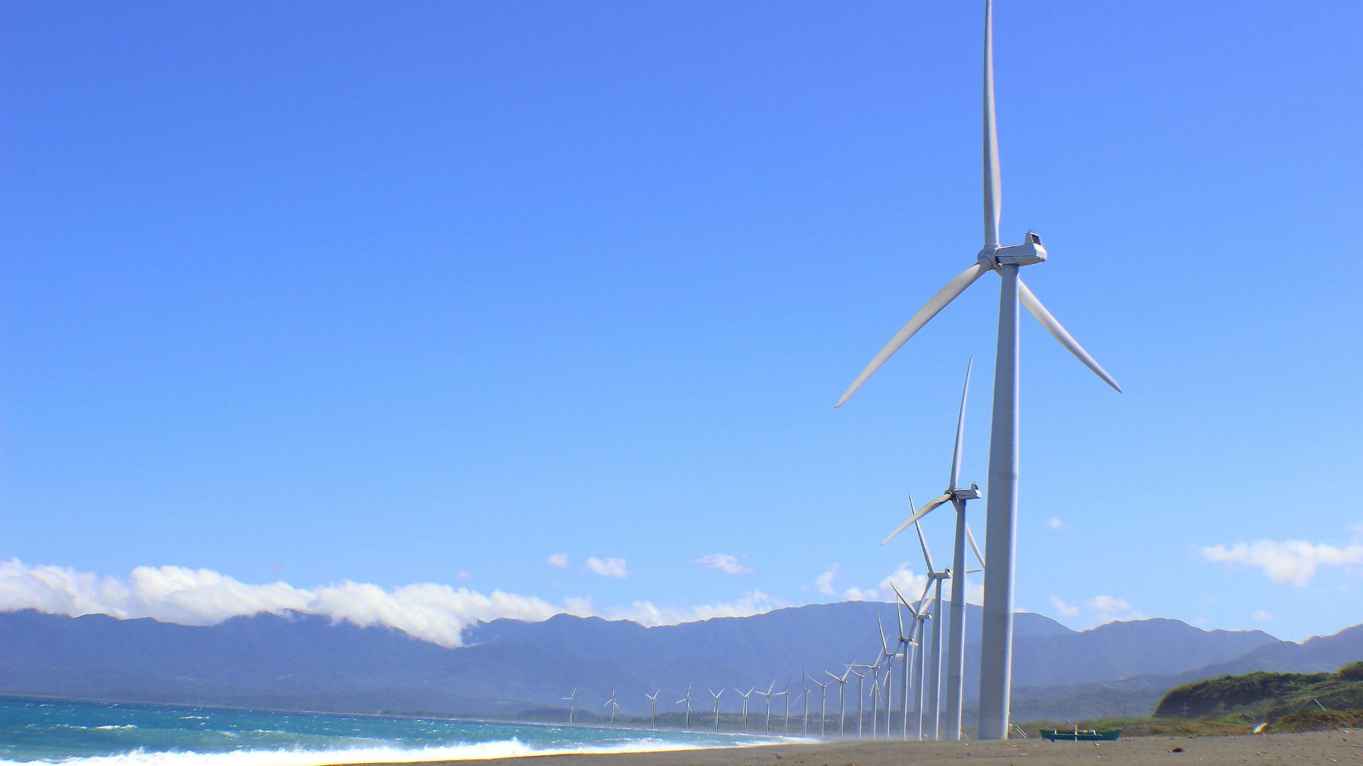 A row of wind turbines next to the ocean extend into the distance. The sky is blue and there are mountains in the distance. 
