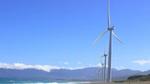 A row of wind turbines next to the ocean extend into the distance. The sky is blue and there are mountains in the distance.